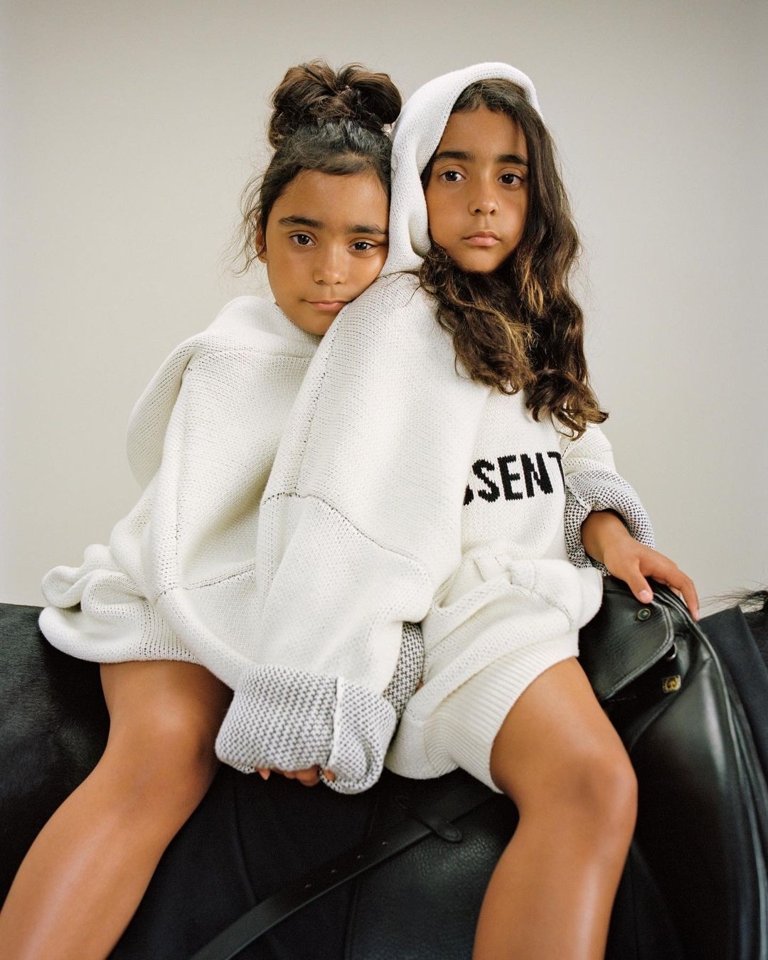 Two young girls in matching knitwear pose cheek-to-cheek while sitting on a black leather horse saddle in a bright, neutral studio.
