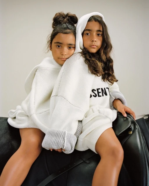 Two young girls in matching knitwear pose cheek-to-cheek while sitting on a black leather horse saddle in a bright, neutral studio.