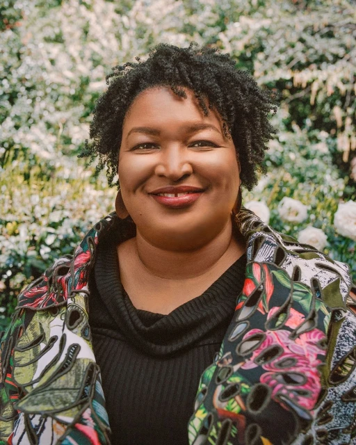 Close-up portrait of an adult woman with curly hair, wearing a patterned jacket, posing in front of a blurred floral background.
