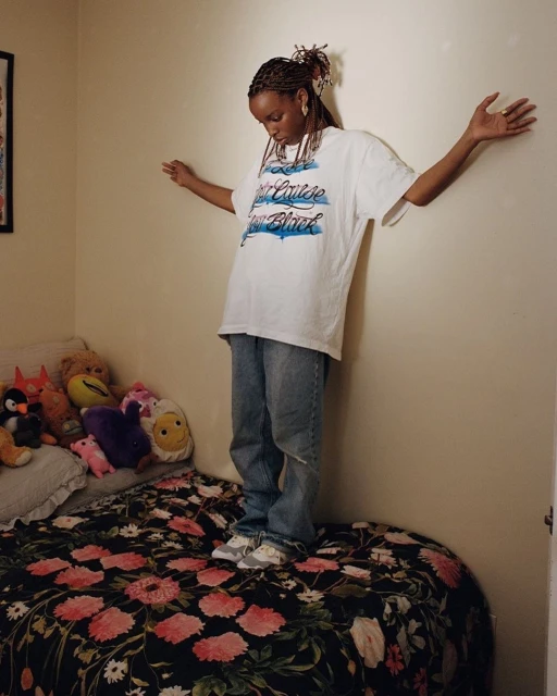 Young woman with braided hair stands on a flower-print bed in an indoor setting, wearing a printed t-shirt and jeans, posing with arms outstretched.