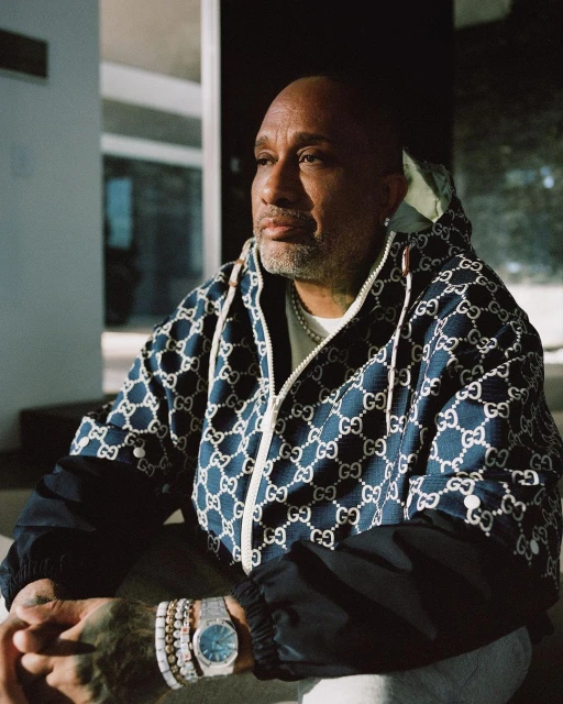 Adult male in a patterned hooded jacket with jewelry poses indoors with a neutral expression, looking off camera in a close-up shot.