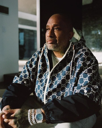 Adult male in a patterned hooded jacket with jewelry poses indoors with a neutral expression, looking off camera in a close-up shot.