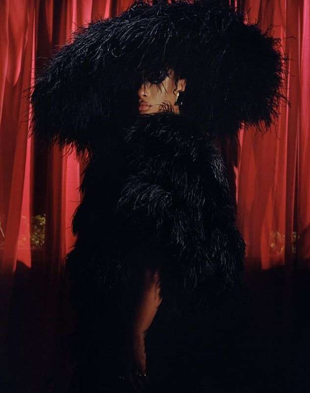 Fashionable adult woman in a large black feather coat and hat, posing against a textured red curtain in a dimly lit indoor setting.