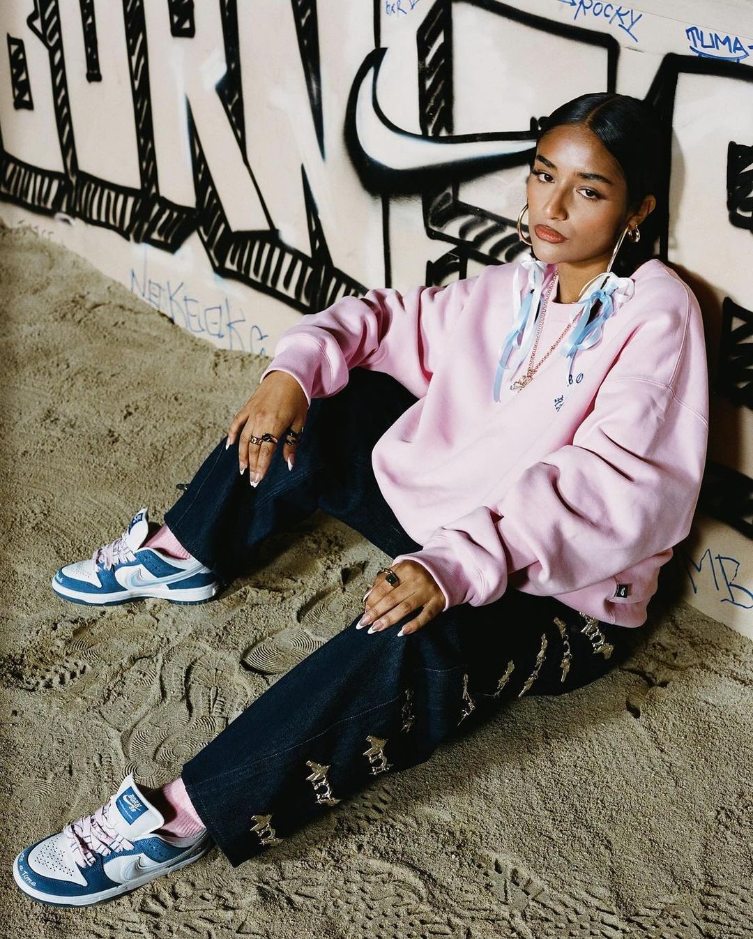 Woman in a pink hoodie and jeans sitting in front of graffiti-covered wall, wearing sneakers and rings in a casual fashion pose.
