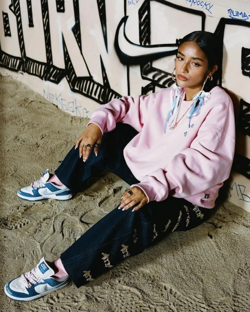 Woman in a pink hoodie and jeans sitting in front of graffiti-covered wall, wearing sneakers and rings in a casual fashion pose.