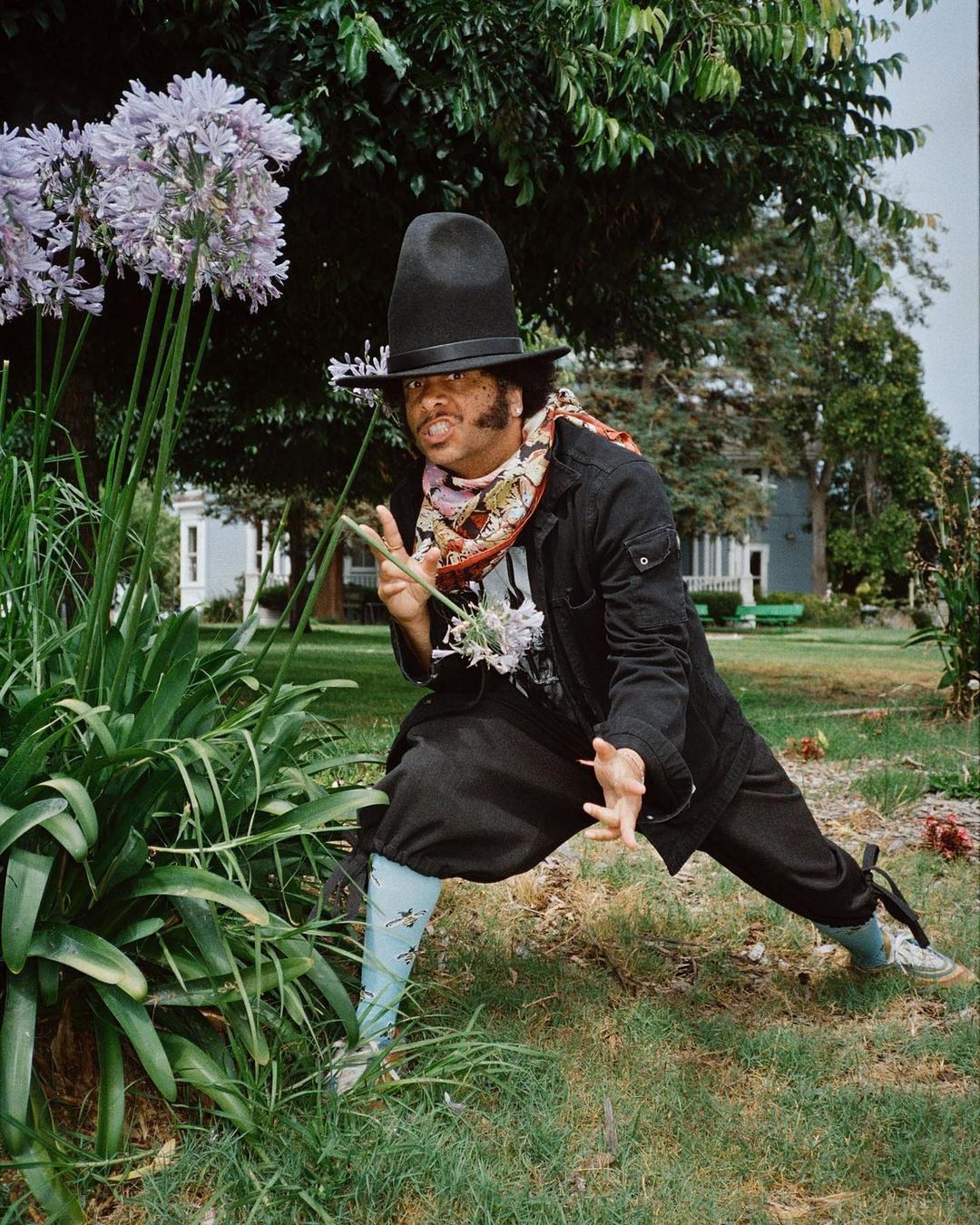Person in a black hat poses in a garden with purple flowers and lush green foliage in an eye-level medium shot during the daytime.