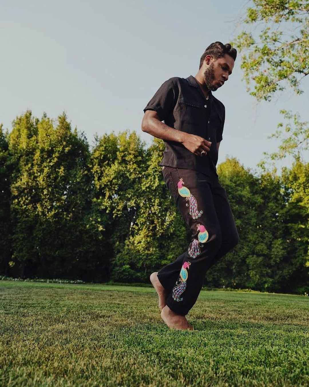Adult male dances barefoot on grass in a black shirt and embroidered pants, with trees in background during daytime.