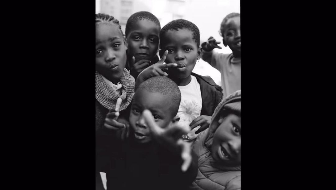 Black and white shot of a group of Black children in an outdoor setting, wearing casual clothing, captured with a close-up, eye-level angle.