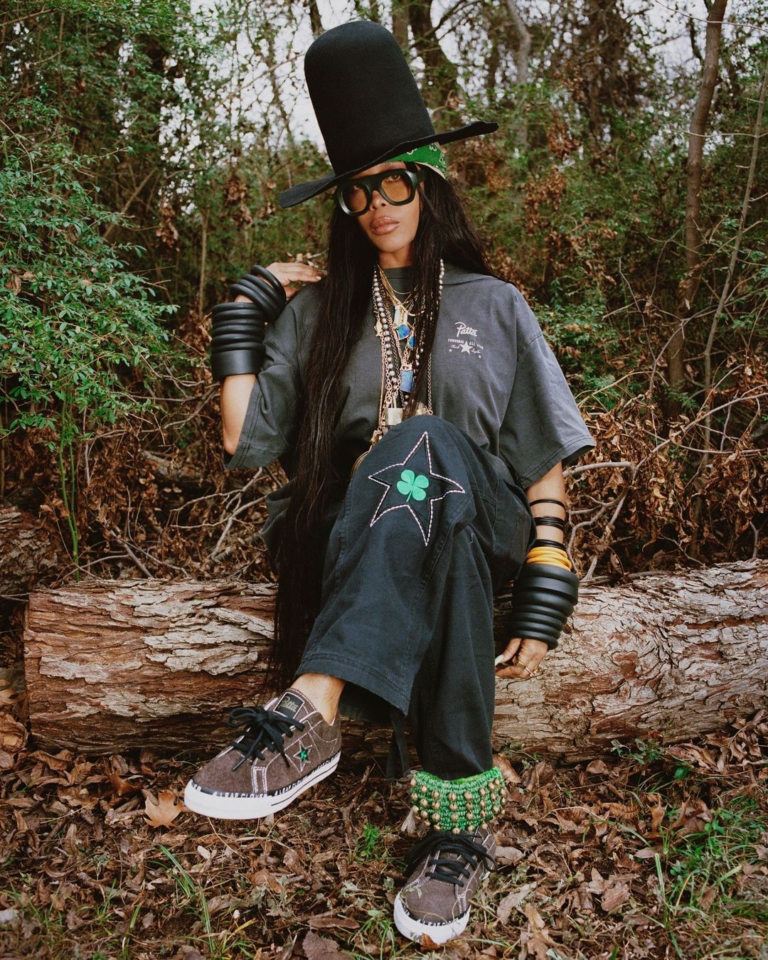 Stylish woman with tall hat sitting on log in nature. She wears sunglasses, bracelets, casual shirt and pants with clover and stars.