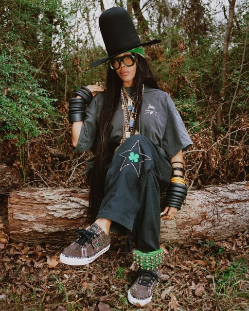 Stylish woman with tall hat sitting on log in nature. She wears sunglasses, bracelets, casual shirt and pants with clover and stars.