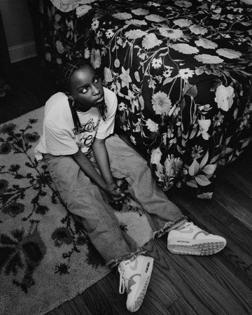 Black and white portrait of a young woman seated on a patterned rug beside a floral bedspread, wearing casual clothes and sneakers.