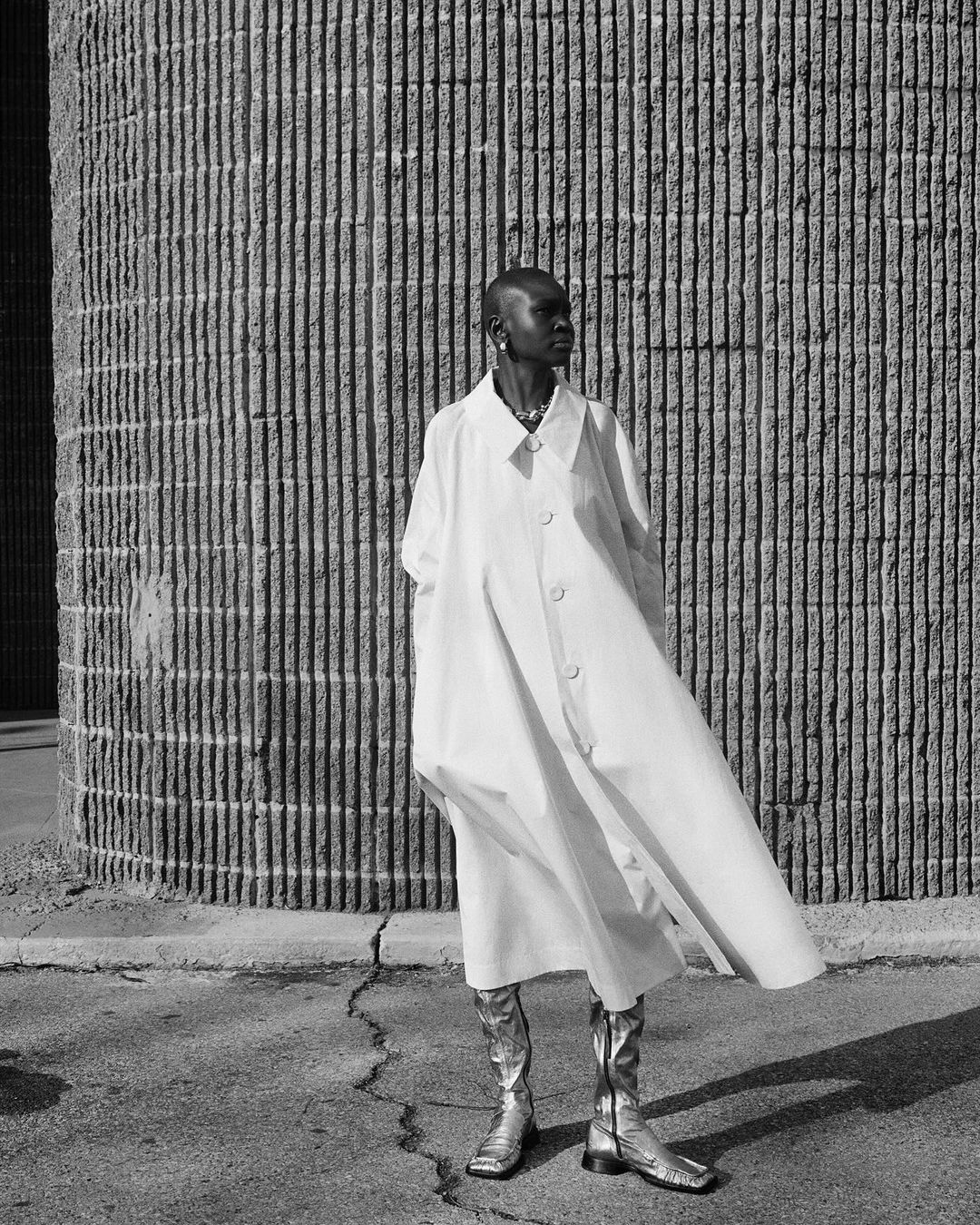 Stylish woman in a white coat and silver boots poses against a textured concrete wall, bathed in soft sunlight, creating a modern urban aesthetic.