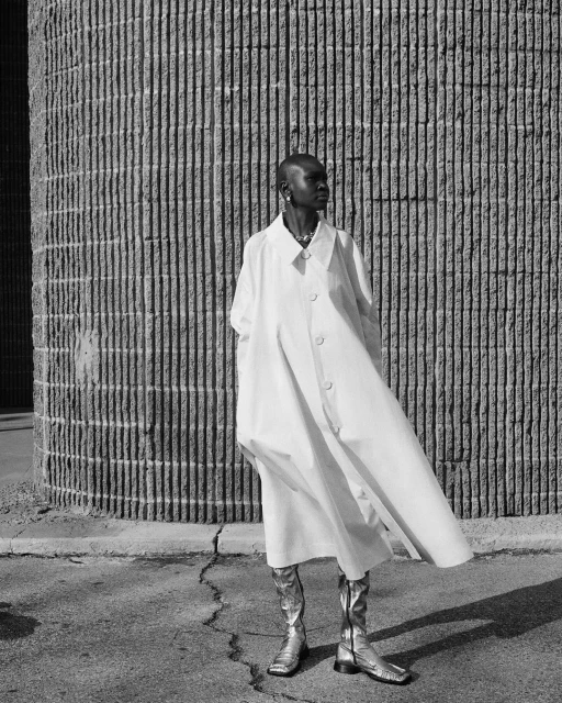 Stylish woman in a white coat and silver boots poses against a textured concrete wall, bathed in soft sunlight, creating a modern urban aesthetic.