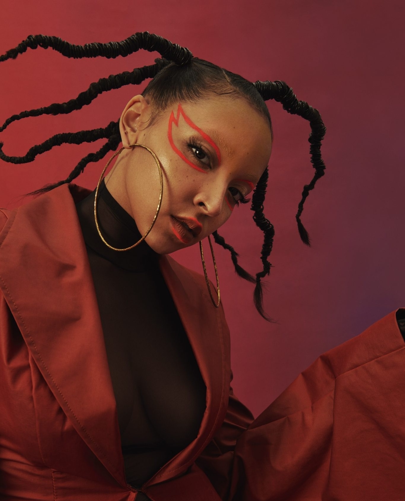 Woman with striking red makeup and distinctive braided hair posing against a gradient red background in a studio setting, wearing gold hoops.
