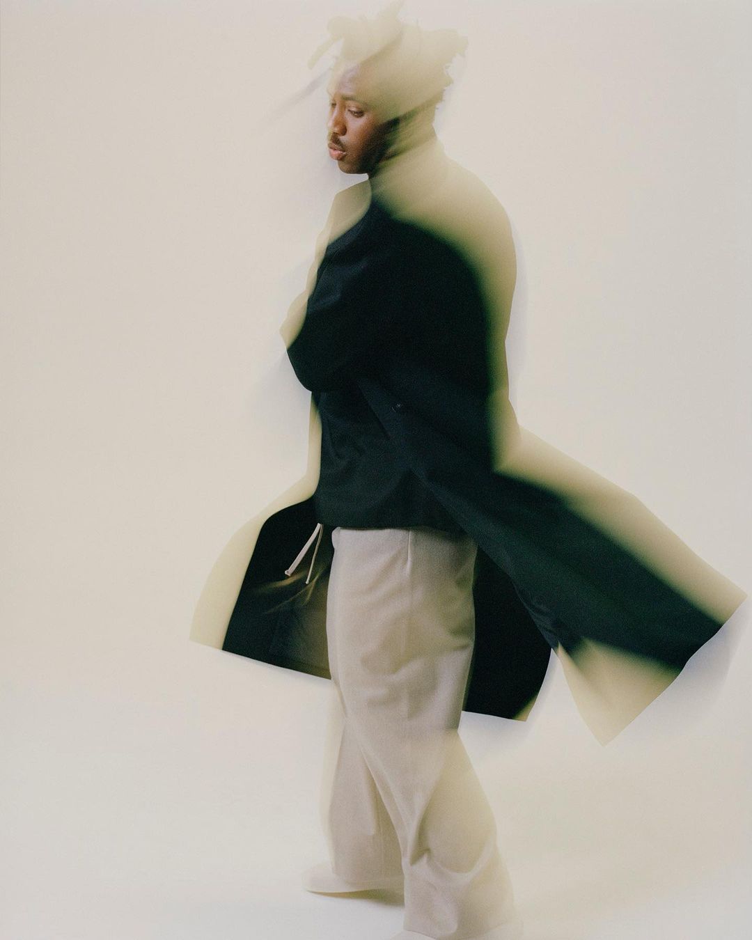 Adult stands in a studio, wearing wide-legged trousers, a black shirt, and a long, dark coat, captured with motion blur against a white backdrop.