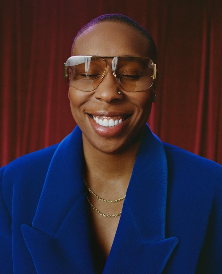 Close-up portrait of a smiling adult female with closed eyes wearing glasses and a blue blazer against a red curtain background.