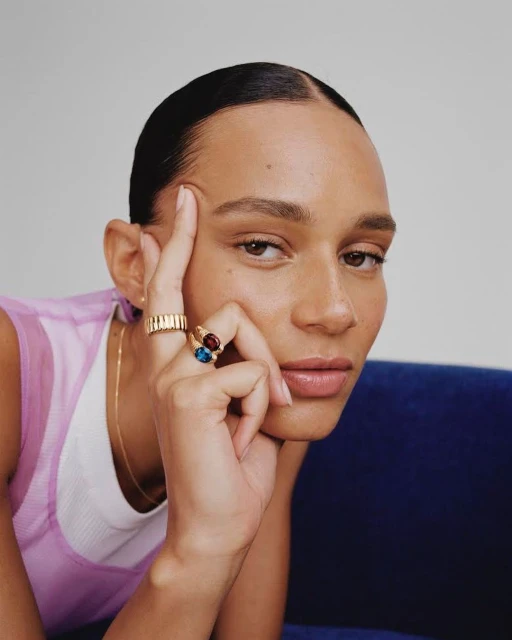 Close-up portrait of a woman with slicked-back dark hair wearing gold rings and a lavender top, resting her chin on her hand indoors.