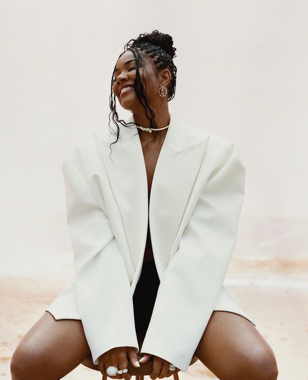 Portrait of a smiling African-American woman wearing an oversized white blazer, statement jewelry, and a hair updo sitting indoors.