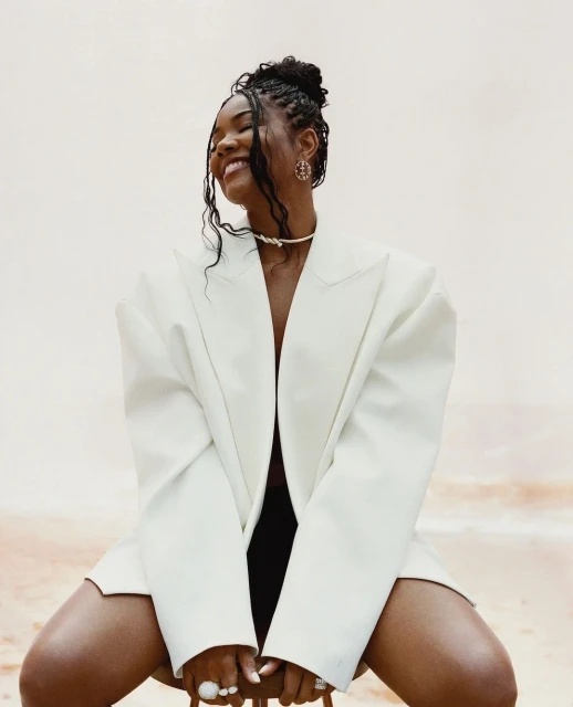 Portrait of a smiling African-American woman wearing an oversized white blazer, statement jewelry, and a hair updo sitting indoors.