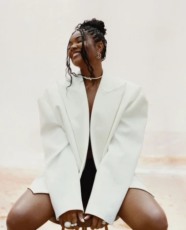 Portrait of a smiling African-American woman wearing an oversized white blazer, statement jewelry, and a hair updo sitting indoors.