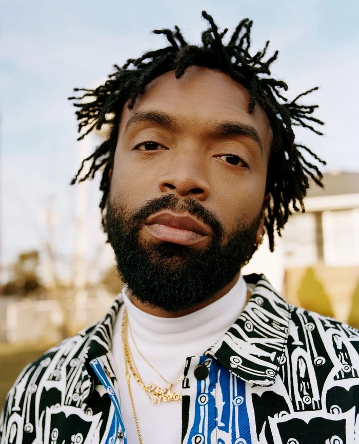 Close-up portrait of a Black man with short dreadlocks wearing a patterned shirt, white turtleneck, and gold necklaces.