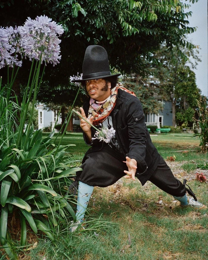 Man in black top hat and jacket posing near tall purple flowers outdoors by Shaniqwa Jarvis