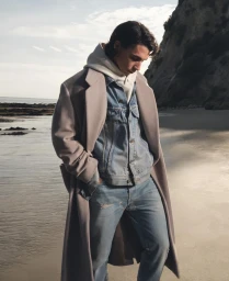 Young man in long camel coat, denim jacket, and jeans standing on sandy beach