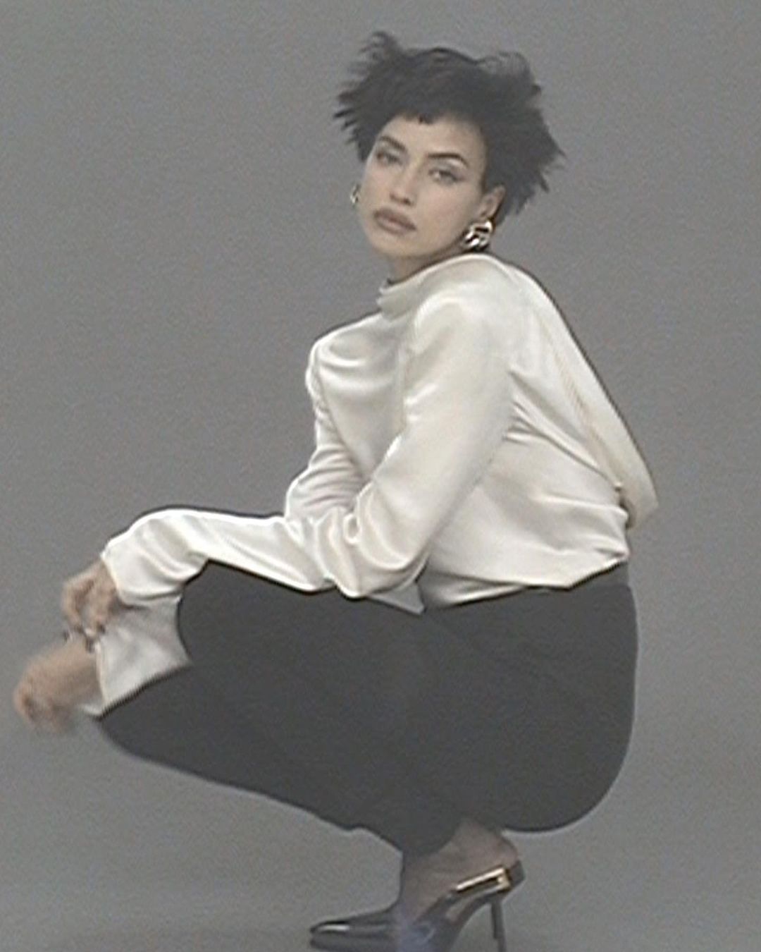 An adult female with short dark hair, wearing a white blouse and black pants, is crouching indoors in front of a solid grey background.