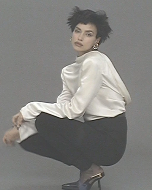 An adult female with short dark hair, wearing a white blouse and black pants, is crouching indoors in front of a solid grey background.