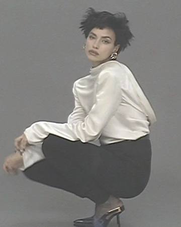An adult female with short dark hair, wearing a white blouse and black pants, is crouching indoors in front of a solid grey background.