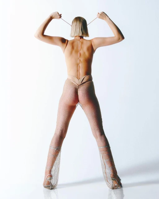 Back view of a slender adult female with bobbed blonde hair raising a drawstring and wearing mesh pants in a bright photo studio.