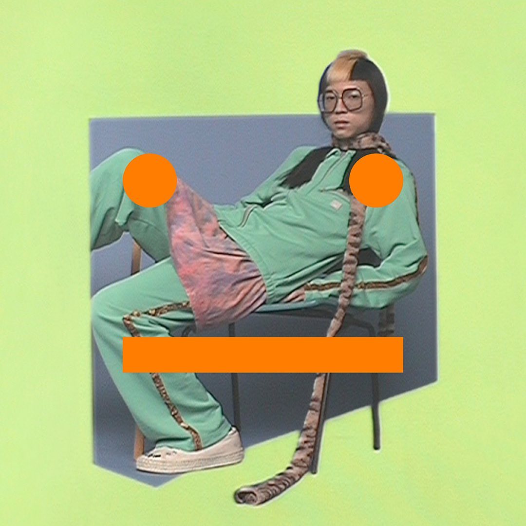 Person in green tracksuit and glasses sits in a chair against a blue and green backdrop in a creative studio portrait.