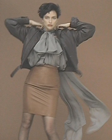 Model with dark hair wearing a dark leather jacket over a grey blouse and tan skirt against a textured brown background