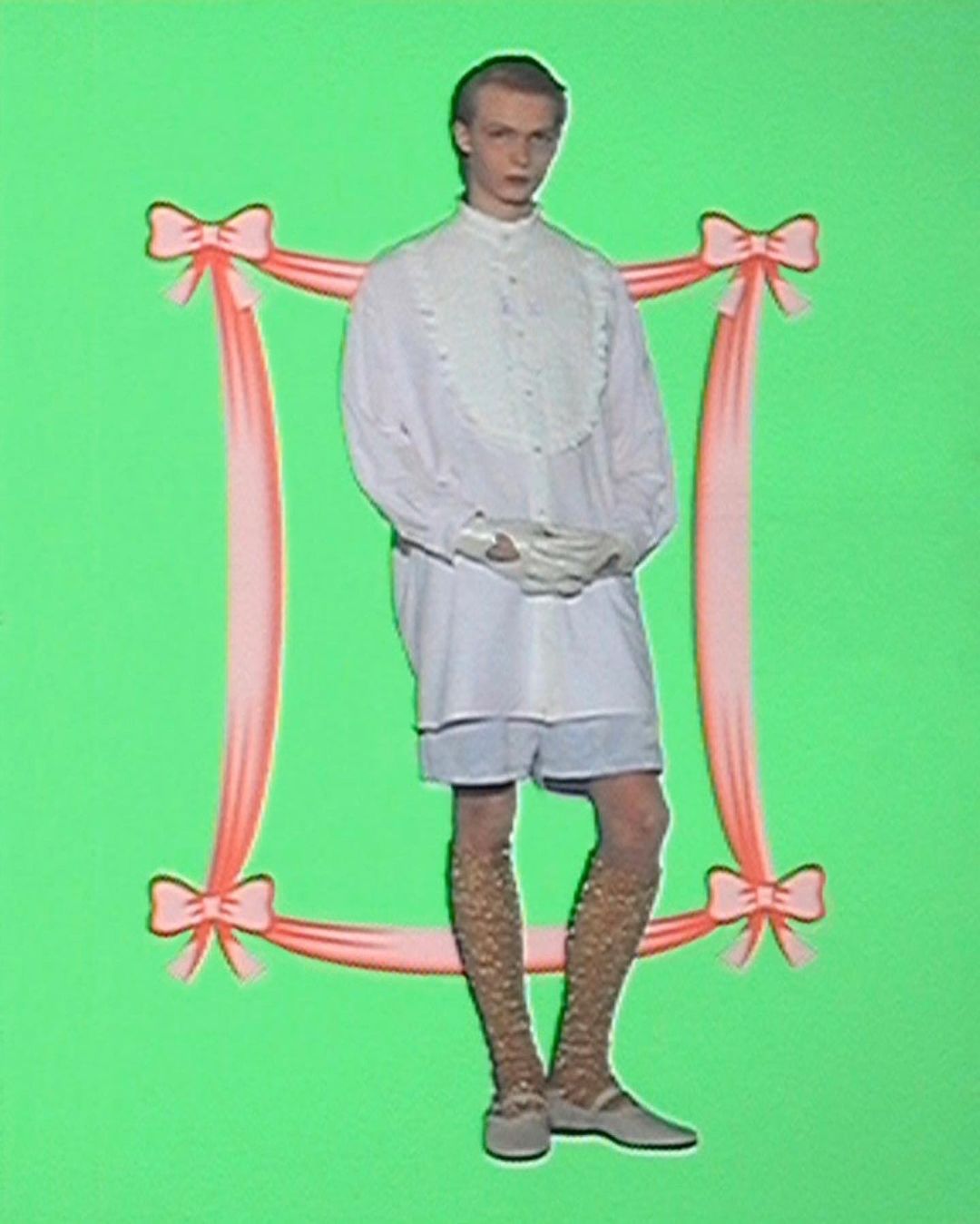 A person poses against a green backdrop, framed by pink ribbon, showcasing a white collared shirt, shorts, patterned stockings, and flats.