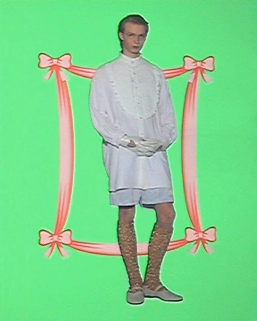 A person poses against a green backdrop, framed by pink ribbon, showcasing a white collared shirt, shorts, patterned stockings, and flats.