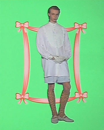 A person poses against a green backdrop, framed by pink ribbon, showcasing a white collared shirt, shorts, patterned stockings, and flats.