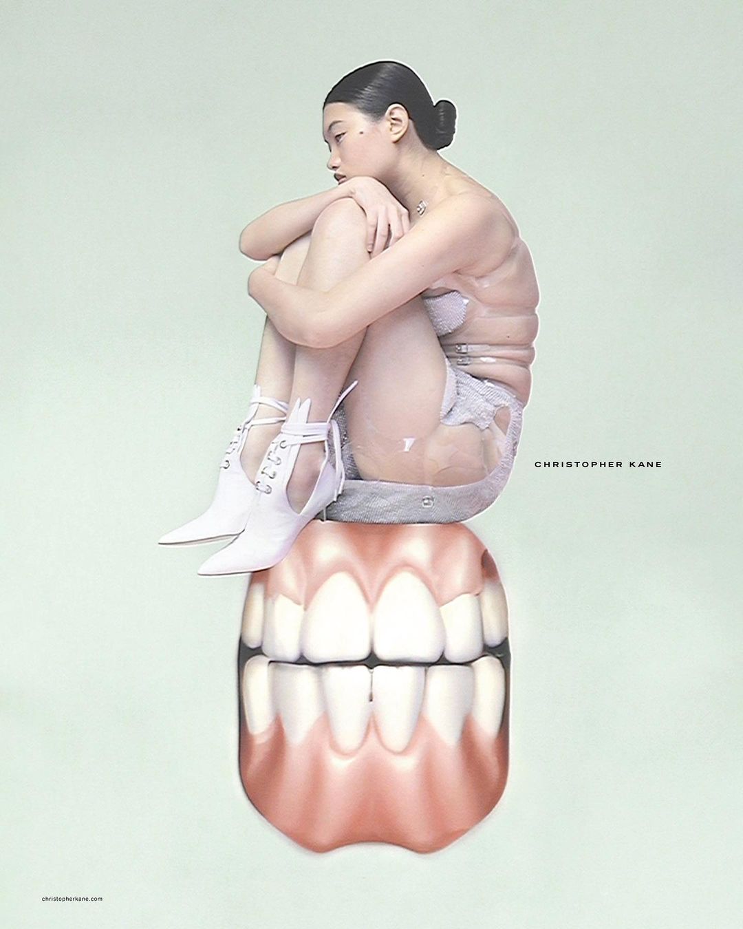 An Asian female model in lingerie and white boots sits atop a large set of teeth in a surreal fashion shot with mint green background.