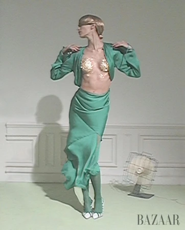 Fashion model in a green ensemble poses in a studio with gold details, complemented by an antique fan and Bazaar logo.