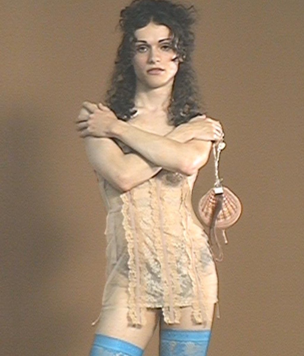 Adult wearing lace lingerie, blue thigh-high stockings, and holding a seashell purse posing in front of a neutral brown backdrop.