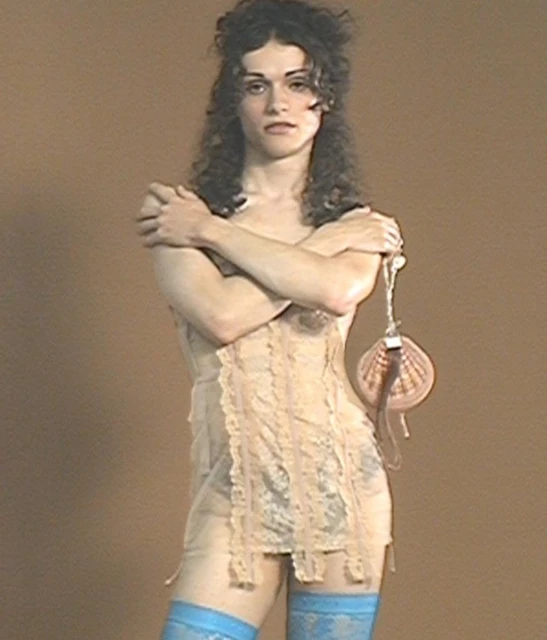 Adult wearing lace lingerie, blue thigh-high stockings, and holding a seashell purse posing in front of a neutral brown backdrop.