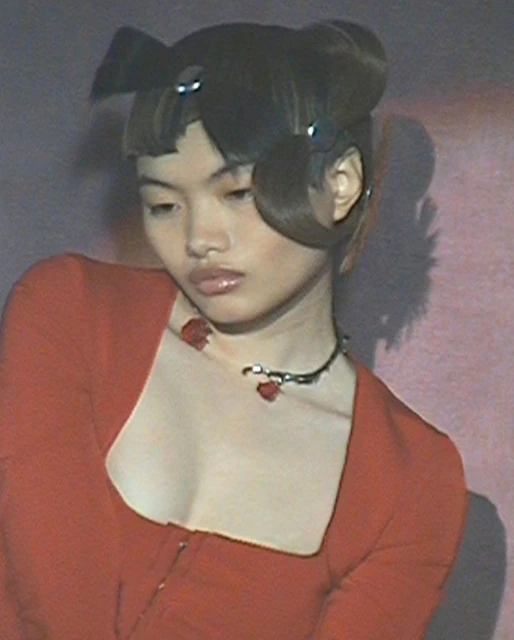 Young woman with stylized hair poses in a close-up shot. She wears a red dress and a necklace with a floral design against a pink backdrop.