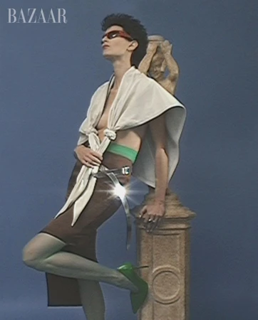 A woman poses in a brown skirt, green stockings and lime heels, next to a sculpture in a studio with a blue backdrop and 'BAZAAR' in corner.
