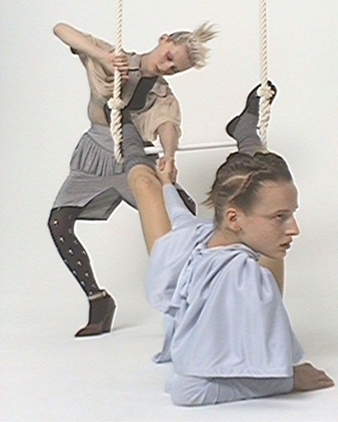 Two fair-skinned young women perform an acrobatic pose on a white background while holding onto white ropes. One woman is above, one below.
