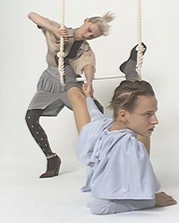 Two fair-skinned young women perform an acrobatic pose on a white background while holding onto white ropes. One woman is above, one below.
