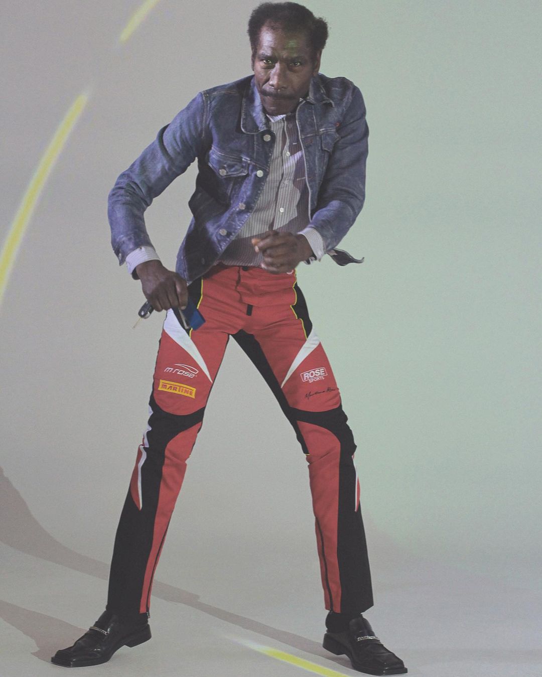 Adult male standing indoors wearing a denim jacket, striped shirt, and racing pants, captured in a studio with dynamic lighting.