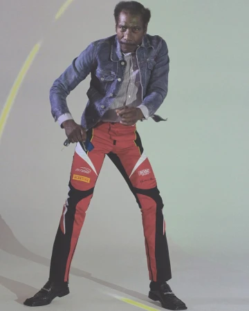 Adult male standing indoors wearing a denim jacket, striped shirt, and racing pants, captured in a studio with dynamic lighting.