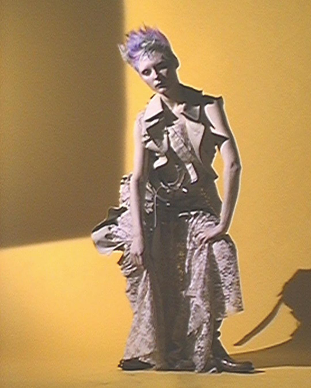 Woman with purple hair in vest and lace skirt poses against yellow background in studio lighting. Fashion editorial photography.