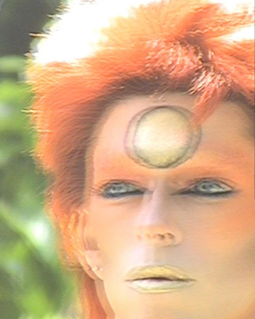 Close-up portrait of a person with orange hair, face makeup, and a painted circle on their forehead, set against a soft green background.