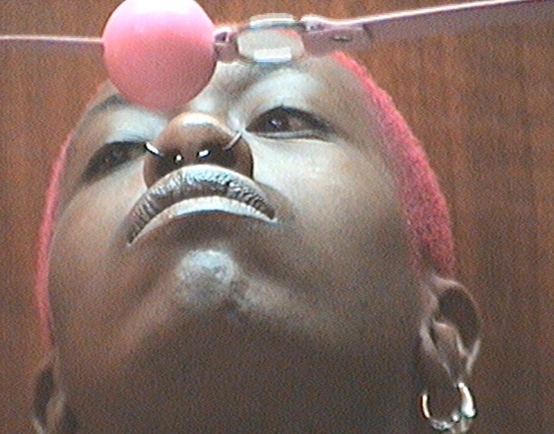 Close-up of a Black adult female with short pink hair looking up, wearing hoop earrings and a nose ring, with a pink ball above her head.