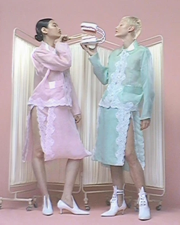 Two models in translucent lace-trimmed outfits strike poses in a pastel photo studio holding dental prop in a pastel pink setting.